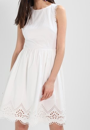 Vrouw draagt een mouwloze witte jurk met een gegolfde zoom en bloemenachtige uitsnijdingen, staande tegen een effen lichte achtergrond.