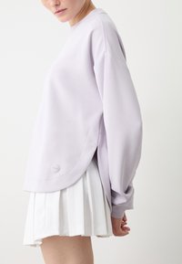 Felpa oversize lavanda con scollo rotondo, spalle cadenti e spacchi laterali, abbinata a una gonna plissettata bianca.