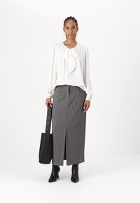 Blusa bianca con fiocco, yoke strutturato; gonna maxi grigia a righe con spacco laterale; stivali neri con tacco; borsa a tracolla nera.