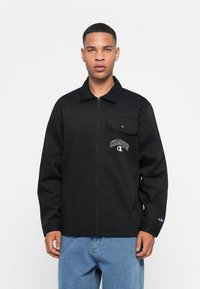Svart zip-up jakke med kragehals, brystlomme og "Champion" logo detalj. Brukt med blå denimbukser, glatt tekstur.