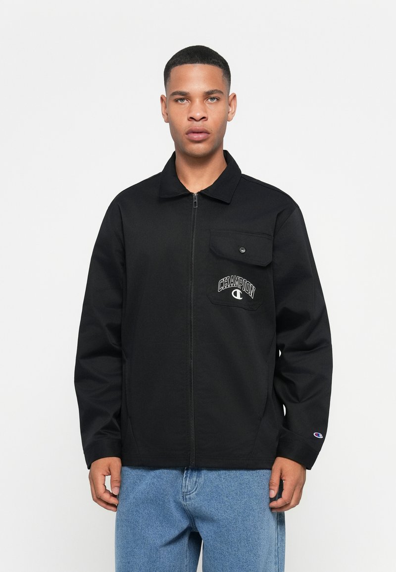 Svart zip-up jakke med kragehals, brystlomme og "Champion" logo detalj. Brukt med blå denimbukser, glatt tekstur.