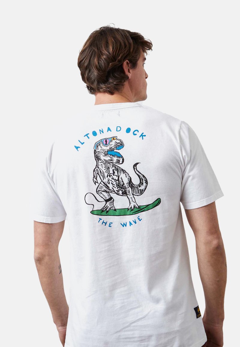 Camiseta de algodón blanca con un gráfico colorido de un dinosaurio en la espalda, un diseño de tabla de surf y un texto que dice "ALTONADOCK THE WAVE".