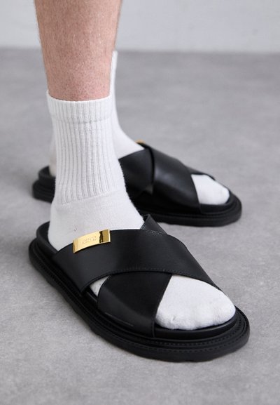 Person som bär svarta läder sandaler med korsremmar över vita ribbade strumpor, står på ett grått golv.