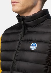 Veste matelassée noire avec un col montant, fermeture éclair à l'avant et logo North Sails sur la poitrine. Présente une texture matelassée et des panneaux latéraux jaunes.