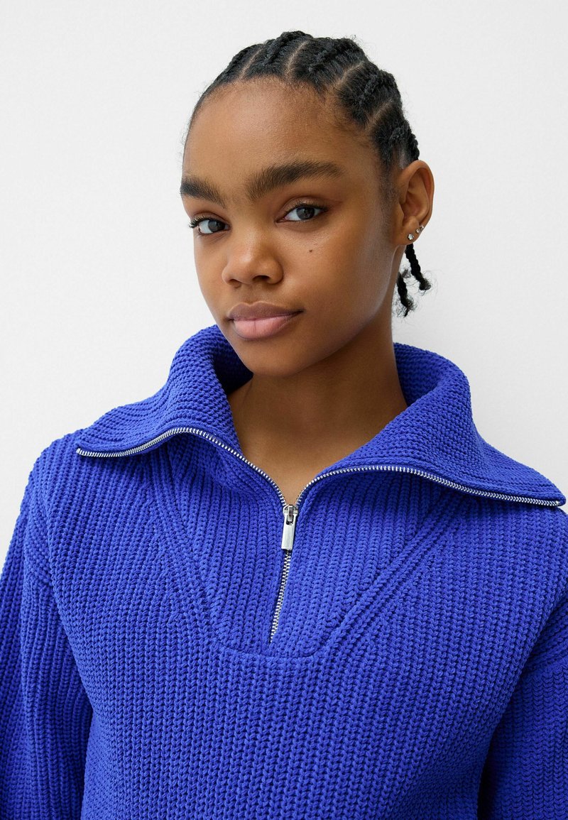 Giovane donna con capelli intrecciati che indossa un pullover lavorato a maglia blu cobalto con una cerniera argentata e colletto ampio, su uno sfondo bianco.