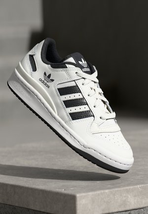 Baskets blanches Adidas avec des bandes et des lacets noirs, flottant au-dessus d'une surface en pierre sur un fond sombre et flou.