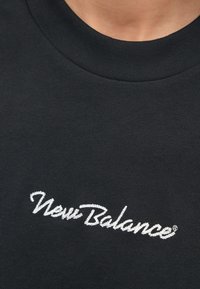 Enfoque próximo de um colarinho de camisa preta com o logótipo "New Balance" em branco bordado centrado no peito.