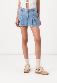 Lichtblauwe spijker mini-jurk met geplooid ontwerp, voorzien van een knoopsluiting en riemlussen. Gecombineerd met beige en bruine sneakers met patroon.