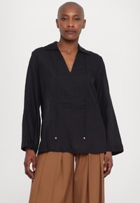 Blusa in lino nera con scollo a V, maniche lunghe e cordoncini con dettagli dorati. Abbinata a pantaloni larghi marroni.