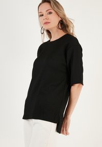 Black short-sleeve top with a round neck, ribbed texture, and side slits, paired with white pants; features intricate hoop earrings.