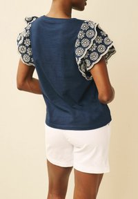 Person wearing navy blue top with white floral lace ruffle sleeves and white shorts, standing against a beige background, viewed from the back.