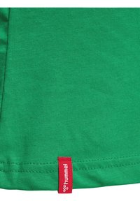 T-shirt en coton vert avec une étiquette de marque rouge à l'ourlet. Le tissu a une texture lisse avec une coupe régulière et sans imprimés visibles.
