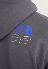 Sudadera gris con un cuello alto, con texto y logo impresos en azul y blanco en el pecho. La tela parece suave con una textura lisa.