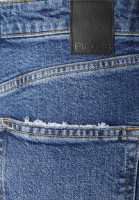 Jeans de ganga azul com um patch de couro preto com a palavra "Pieces", costura amarela e bordas desfiadas ao longo da bainha e dos bolsos.