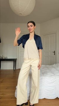 Νέα γυναίκα με σκούρα μαλλιά σε λευκό τοπ με δαντέλα, σκούρο μπλε κοντό σακάκι, κρεμ παντελόνι φαρδιάς γραμμής και μαύρα παπούτσια που χαιρετά σε υπνοδωμάτιο.