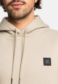 Beige hoodie met een trekkoord, voorzien van een zwart vierkant logo-opdruk aan de voorkant. De stof lijkt zacht en textuur te hebben.