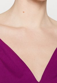 Bluzka z dekoltem w serek w głębokim fioletowym materiale, z gładką fakturą i starannym ściem wzdłuż krawędzi, podkreślającym linię dekoltu i ramion.