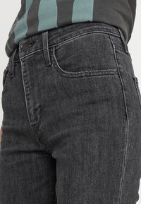 Jeans gris oscuro con corte recto, diseño de cinco bolsillos, costuras visibles y cierre de botón en la cintura. Textura de denim suave.