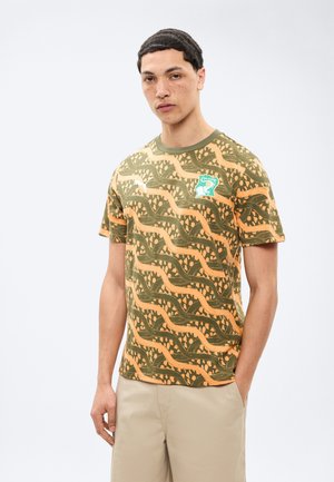 Άνδρας που φοράει πράσινο και πορτοκαλί μπλουζάκι με κυματιστό σχέδιο και μπεζ σορτσάκι, στέκεται με το ένα χέρι στην τσέπη του μπροστά σε λευκό φόντο.