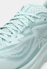 Világoskék futócipő hálós felsőrésszel, lekerekített orral és vastag talppal. Texturált mintázat és Hoka márkajelzés található az oldalán.