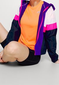 Pestrý sportovní outfit skládající se z oranžového trička, fialové a růžové bundy s bílými detaily a černých kraťasů. Hladké, lehké materiály.