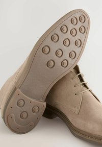 Beige Wildlederschuhe mit runder Zehenpartie und Schnürdesign. Die Gummisohle verfügt über runde Traktionsmuster und ein lächelndes Gesicht Detail.