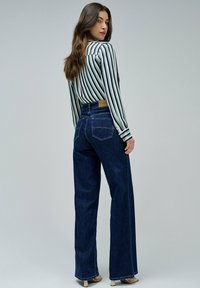 Jean en denim sombre à jambes larges associé à une chemise rayée verte et blanche. Le tissu a une texture lisse, avec des détails de couture visibles.