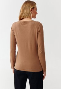Bluza lunga maro cu mâneci, confecționată din material moale, cu guler rotunjit și siluetă ajustată, asortată cu pantaloni închiși la culoare. Fără modele vizibile.