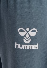 Grijze stof met een gestructureerde weving, met een wit logo van gestileerde vleugels en een blad, prominent gepositioneerd aan de onderkant.