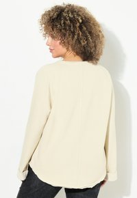 Top de manga larga color crema con dobladillo redondeado y puños enrollados. Tejido suave con un ajuste relajado y mangas raglán.
