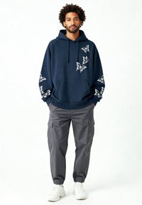 Sweat à capuche bleu marine avec broderie de papillons blancs sur les manches et la poche avant ; assorti à un pantalon cargo gris et des baskets blanches. Coupe décontractée.