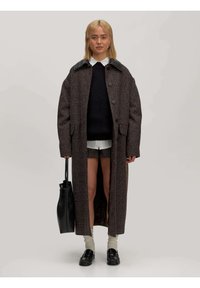 Langer, strukturierter brauner Mantel mit schwarzem Lederkragen, mit einem einzigen Knopfverschluss, über einem dunklen Pullover und Shorts getragen. Schwarze Tasche und Schuhe, cremefarbene Socken.