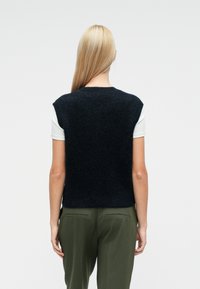 Πλεκτή αμάνικη μπλούζα σε σκούρο ναυτικό μπλε με στρογγυλή λαιμόκοψη, φορεμένη πάνω από ένα λευκό πουκάμισο, σε συνδυασμό με χακί πράσινο παντελόνι. Λείο υφής.