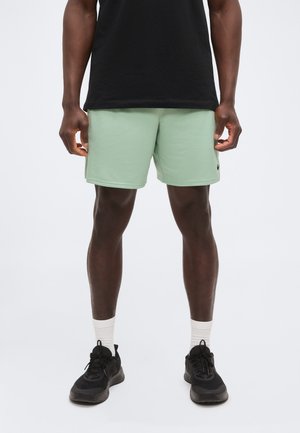 Homme portant un short vert clair, des baskets noires, des chaussettes blanches et un t-shirt noir, debout les jambes légèrement écartées sur un fond uni.