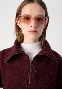 Bordeaux zip-up trui met een hoge kraag, gestructureerde brei en een gouden ritssluiting; gecombineerd met transparante roze zonnebril.