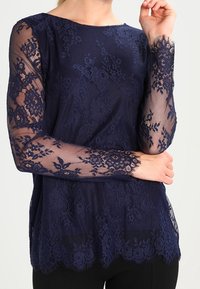 Top a maniche lunghe blu navy realizzata in pizzo, con motivi floreali e finitura trasparente sulle maniche, dotato di orlo smerlato.