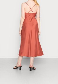 Satinové midi šaty v spálené oranžové barvě s volným střihem. Mají tenké křížové ramínka a hladkou texturu. Nosené s černými sandály na blokovém podpatku.