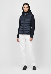 Granatowa pikowana kamizelka z kapturem, długie rękawy z prążkowanym wykończeniem, zapięcie na zamek błyskawiczny i pikowana faktura, zestawiona z białymi spodniami i czarnymi butami.