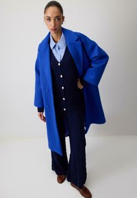 Lang blå frakke i tykk stof, lagdelt over en marineblå kabelstrikvest med store knapper, parret med matchende strikkede bukser og brune sko.