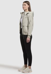 Light beige hooded jacket with multiple pockets, zip closure, and textured fabric, paired with black leggings and light sneakers.