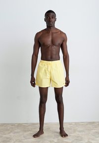 Un short de bain jaune avec une taille élastique et un cordon de serrage, fabriqué en tissu léger, présenté sur un modèle avec un arrière-plan neutre.