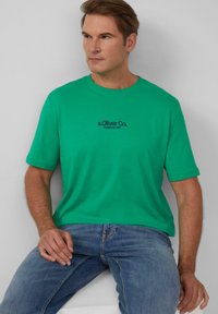 Zielona bawełniana koszulka z krótkim rękawem i okrągłym dekoltem, z czarnym nadrukowanym logo na piersi. W połączeniu z niebieskimi dżinsami.