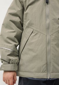 Olijfgroene jas met een volledige rits, elastische manchetten en reflecterende accenten. Het materiaal heeft een gladde textuur en een iets gestructureerd ontwerp.