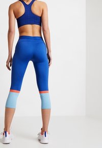 Modré sportovní legíny s texturovaným světle modrým lemováním, oranžovými akcenty a délkou po lýtka. Doplněné odpovídajícím modrým sportovním braletem.