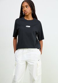Crna majica s kratkim rukavima s bijelim Nike logom na prsima. Uparena s bijelim cargo hlačama koje imaju bočne džepove. Minimalistički dizajn.