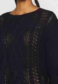 Pull en maille bleu marine présentant un motif texturé, des sections à mailles ouvertes et un large col, mettant en valeur un design détaillé en tricot torsadé.