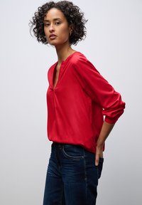 Červená blůza s dlouhými rukávy a výstřihem do V, vyrobená z hladkého materiálu. V kombinaci s tmavě modrými džíny. Jednoduchý střih bez výrazných vzorů nebo akcentů.