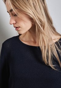 Marineblauwe, gebreide trui met textuur en een brede halslijn, met lange mouwen en een ontspannen pasvorm, gedragen door een persoon met lichtbruin haar.