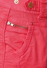 Red cotton pants featuring a zippered pocket, decorative stitching, and a metal logo accent on the waistband.