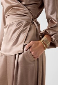 Top avvolgente in satin beige con maniche lunghe e nodo laterale. Caratterizzato da una morbida texture e dettagli plissettati, completato da un braccialetto a manica dorato.